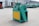 A green and yellow industrial machine is positioned outdoors on a concrete surface. The machine has a large belt and is situated in front of a building with a corrugated metal exterior. Other industrial equipment is visible in the background.