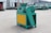 A green and yellow industrial machine is positioned outdoors on a concrete surface. The machine has a large belt and is situated in front of a building with a corrugated metal exterior. Other industrial equipment is visible in the background.