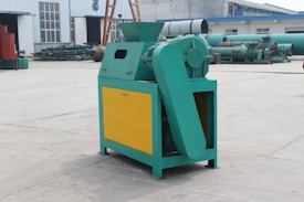 A green and yellow industrial machine is positioned outdoors on a concrete surface. The machine has a large belt and is situated in front of a building with a corrugated metal exterior. Other industrial equipment is visible in the background.