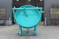 A massive teal-colored industrial drum with a flat top stands in an exterior factory setting. The structure is placed on a flat concrete surface, flanked by gray metal walls with windows and some industrial piping. Scaffolding is visible on the right side.