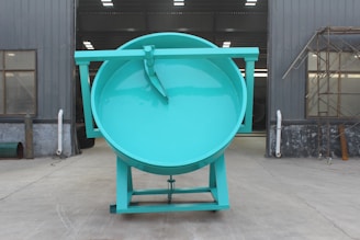 A massive teal-colored industrial drum with a flat top stands in an exterior factory setting. The structure is placed on a flat concrete surface, flanked by gray metal walls with windows and some industrial piping. Scaffolding is visible on the right side.