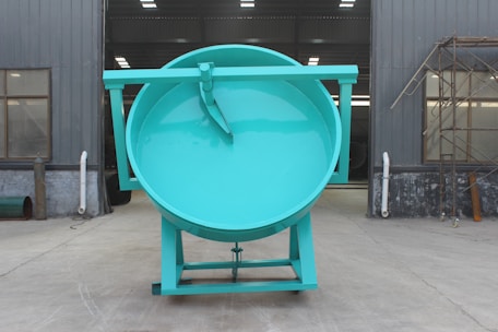 A massive teal-colored industrial drum with a flat top stands in an exterior factory setting. The structure is placed on a flat concrete surface, flanked by gray metal walls with windows and some industrial piping. Scaffolding is visible on the right side.