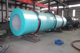 A large industrial cylindrical machine wrapped in a turquoise covering is positioned in a spacious factory setting with high ceilings and overhead beams. The machinery has visible gears and is mounted on a sturdy base. Nearby on the floor, there is a blue motor and various pieces of metal equipment scattered around. The industrial environment is well-lit, with natural light coming from the windows above.