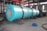 A large industrial cylindrical machine wrapped in a turquoise covering is positioned in a spacious factory setting with high ceilings and overhead beams. The machinery has visible gears and is mounted on a sturdy base. Nearby on the floor, there is a blue motor and various pieces of metal equipment scattered around. The industrial environment is well-lit, with natural light coming from the windows above.