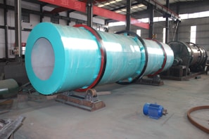 A large industrial cylindrical machine wrapped in a turquoise covering is positioned in a spacious factory setting with high ceilings and overhead beams. The machinery has visible gears and is mounted on a sturdy base. Nearby on the floor, there is a blue motor and various pieces of metal equipment scattered around. The industrial environment is well-lit, with natural light coming from the windows above.