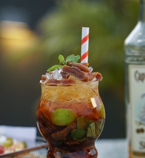 A glass of refreshing cocktail garnished with mint leaves, lime slices, and possibly figs, with ice. The drink has a swirling pattern of dark and light brown layers, and is topped with a white and red striped straw. Beside the drink, a bottle with an obscured label stands slightly out of focus.