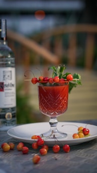 A cocktail glass filled with a red beverage is garnished with cherries on skewers and fresh herbs. The setup includes a white plate with scattered cherries and a bottle of vodka to the side. The background features a blurred garden setting, giving a relaxed outdoor vibe.