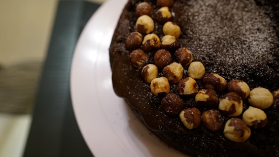 A chocolate cake is partially visible, decorated with a ring of whole hazelnuts and a dusting of powdered sugar on top. The cake appears rich and dark, with a glossy finish on the icing.