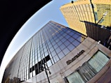 A dramatic upward perspective of a modern skyscraper with reflective glass windows dominates the view. The building, labeled with the name Sheraton, exhibits clean architectural lines and mirrors the blue sky. Adjacent to it, another towering structure features a grid-like design. Street lamps and a street sign reading 'Court' add urban elements to the scene.