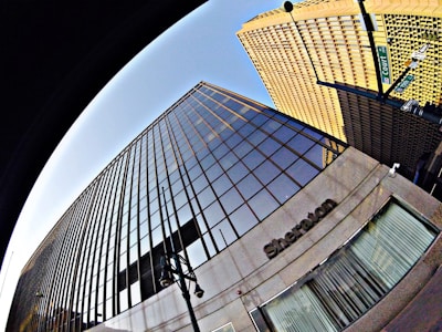 A dramatic upward perspective of a modern skyscraper with reflective glass windows dominates the view. The building, labeled with the name Sheraton, exhibits clean architectural lines and mirrors the blue sky. Adjacent to it, another towering structure features a grid-like design. Street lamps and a street sign reading 'Court' add urban elements to the scene.