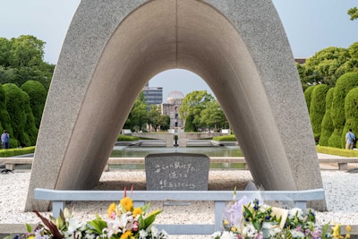 A prominent stone arch monument frames a cenotaph and large memorial in the distance. Floral tributes are placed at the forefront on a bed of pebbles. The scene is set in a tranquil park, surrounded by green, manicured trees and a clear, bright sky.