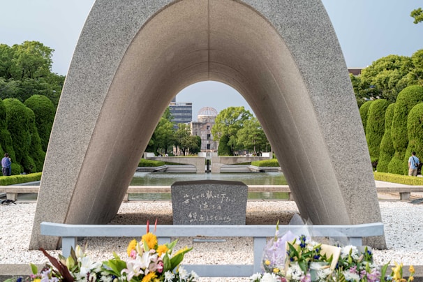 A prominent stone arch monument frames a cenotaph and large memorial in the distance. Floral tributes are placed at the forefront on a bed of pebbles. The scene is set in a tranquil park, surrounded by green, manicured trees and a clear, bright sky.