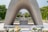 A prominent stone arch monument frames a cenotaph and large memorial in the distance. Floral tributes are placed at the forefront on a bed of pebbles. The scene is set in a tranquil park, surrounded by green, manicured trees and a clear, bright sky.