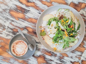 A beautifully arranged salad with slices of apple, leafy greens, and scattered edible flowers, served on a round plate. Next to it, a cup of frothy cappuccino is placed on a saucer with a spoon, all set on a rustic wooden table.
