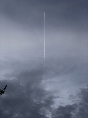 A long, narrow contrail streaks vertically through a cloudy sky, parting the dark and moody atmosphere. The lower portion of the sky features darker clouds, suggesting an overcast or stormy weather condition.