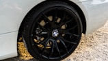 Close-up of a car wheel featuring a black alloy rim with the BMW logo at the center. The car's body is white and the ground is covered in gravel.