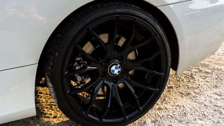 Close-up of a car wheel featuring a black alloy rim with the BMW logo at the center. The car's body is white and the ground is covered in gravel.