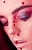 A close-up view of a person's face with red makeup dots scattered across the forehead, cheeks, and around the lips. The eyes are closed, and the eyelids have shiny, multi-colored eyeshadow, primarily pink and purple. The skin has a soft, smooth texture with prominent eyebrows and long lashes.