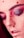 A close-up view of a person's face with red makeup dots scattered across the forehead, cheeks, and around the lips. The eyes are closed, and the eyelids have shiny, multi-colored eyeshadow, primarily pink and purple. The skin has a soft, smooth texture with prominent eyebrows and long lashes.