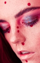 A close-up view of a person's face with red makeup dots scattered across the forehead, cheeks, and around the lips. The eyes are closed, and the eyelids have shiny, multi-colored eyeshadow, primarily pink and purple. The skin has a soft, smooth texture with prominent eyebrows and long lashes.