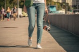 A person walks along a paved area, holding a vintage-style camera in one hand. The person is dressed in casual attire, wearing blue jeans, white sneakers, and a white top. A green shoulder bag is slung across their body. In the background, several people are walking, and some buildings and trees are visible.