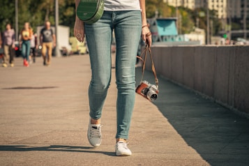 A person walks along a paved area, holding a vintage-style camera in one hand. The person is dressed in casual attire, wearing blue jeans, white sneakers, and a white top. A green shoulder bag is slung across their body. In the background, several people are walking, and some buildings and trees are visible.