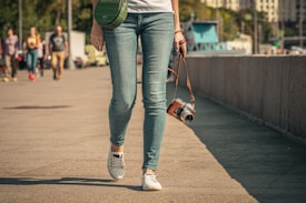 A person walks along a paved area, holding a vintage-style camera in one hand. The person is dressed in casual attire, wearing blue jeans, white sneakers, and a white top. A green shoulder bag is slung across their body. In the background, several people are walking, and some buildings and trees are visible.