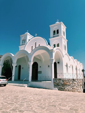 A white, traditional-style church featuring twin bell towers and a series of arches under a bright blue sky. The building is situated on a stone-paved area with a railing around the perimeter. A car is parked nearby, and the structure has multiple arched windows and doors.