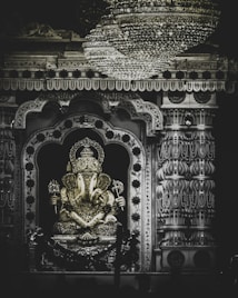 An ornate statue of the Hindu deity Ganesha sits in a beautifully decorated shrine. The intricate details of the statue are highlighted by the numerous jewels and ornaments adorning it. A large, glistening chandelier hangs above, casting light on the richly textured columns and decorative archway surrounding the statue.