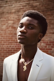 A person wearing a white suit looks confidently at the camera, with a brick wall serving as the backdrop. The individual is adorned with a gold necklace featuring a pendant in the shape of a continent.