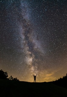 A silhouette of a person standing with arms raised against a backdrop of the Milky Way galaxy in a star-filled night sky. Dark terrain and faint outlines of trees are visible at the bottom, with a warm glow from the horizon.