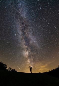 A silhouette of a person standing with arms raised against a backdrop of the Milky Way galaxy in a star-filled night sky. Dark terrain and faint outlines of trees are visible at the bottom, with a warm glow from the horizon.