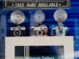 Three vintage cameras with large flash attachments are displayed on a shelf with a sign above them. The sign reads 'TAKE AWAY AVAILABLE' in bold letters. The background is a blurred blue, and there's a reflection of a person in the glass.
