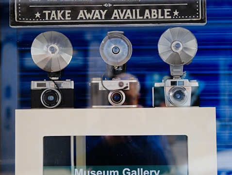 Three vintage cameras with large flash attachments are displayed on a shelf with a sign above them. The sign reads 'TAKE AWAY AVAILABLE' in bold letters. The background is a blurred blue, and there's a reflection of a person in the glass.