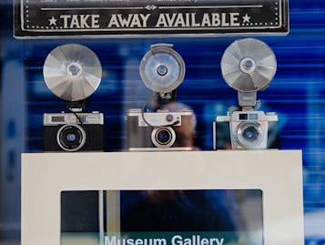 Three vintage cameras with large flash attachments are displayed on a shelf with a sign above them. The sign reads 'TAKE AWAY AVAILABLE' in bold letters. The background is a blurred blue, and there's a reflection of a person in the glass.