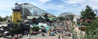 An amusement park scene with a roller coaster and various themed buildings in a bustling environment. Numerous people are walking around the area, which includes greenery, rock formations, and tables with red tops. A lighthouse structure adds a nautical theme to the setting.