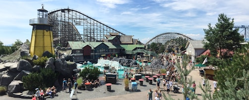 An amusement park scene with a roller coaster and various themed buildings in a bustling environment. Numerous people are walking around the area, which includes greenery, rock formations, and tables with red tops. A lighthouse structure adds a nautical theme to the setting.