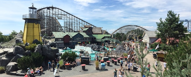 An amusement park scene with a roller coaster and various themed buildings in a bustling environment. Numerous people are walking around the area, which includes greenery, rock formations, and tables with red tops. A lighthouse structure adds a nautical theme to the setting.