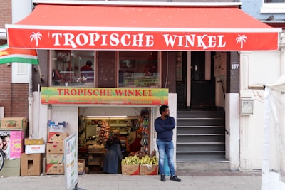 In front of a shop with a red awning displaying the words 'Tropische Winkel', a man stands with his arms crossed next to a fruit stand offering bananas. The store appears to sell Surinamese and Antillean products, evident from the signage. Cardboard boxes are stacked on the left, and there are visible stairs leading to an upper section. A couple of people are inside, attending to the shop.