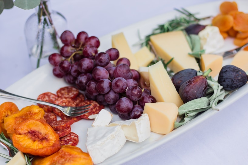 A white platter is arranged with an assortment of cheeses, including wedges and a soft round cheese. Fresh fruits such as red grapes, figs, and apricots accompany the cheese, alongside slices of salami and dried peaches. Fresh herbs like sage add a touch of green to the presentation. The setup suggests an elegant cheese and charcuterie board.