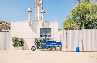 A blue three-wheeled vehicle is parked in front of a light-colored industrial building with two tall metal chimneys. There are some green plants in pots near the vehicle and a larger tree to the right. The area is brightly lit with clear skies, and a tall fence is visible behind the vehicle.