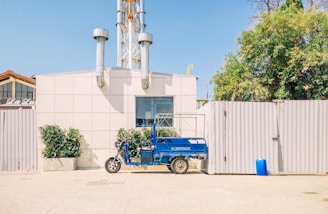 A blue three-wheeled vehicle is parked in front of a light-colored industrial building with two tall metal chimneys. There are some green plants in pots near the vehicle and a larger tree to the right. The area is brightly lit with clear skies, and a tall fence is visible behind the vehicle.