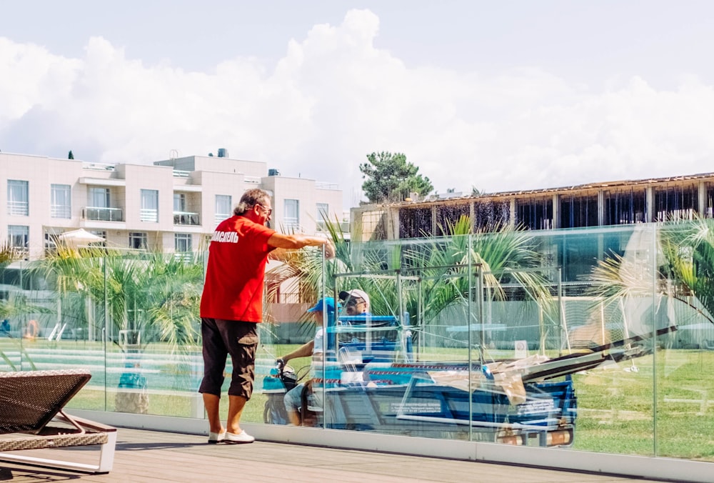 man wearing red and white t-shirt standing near railings and another two person sitting in boat viewing buildings during daytime