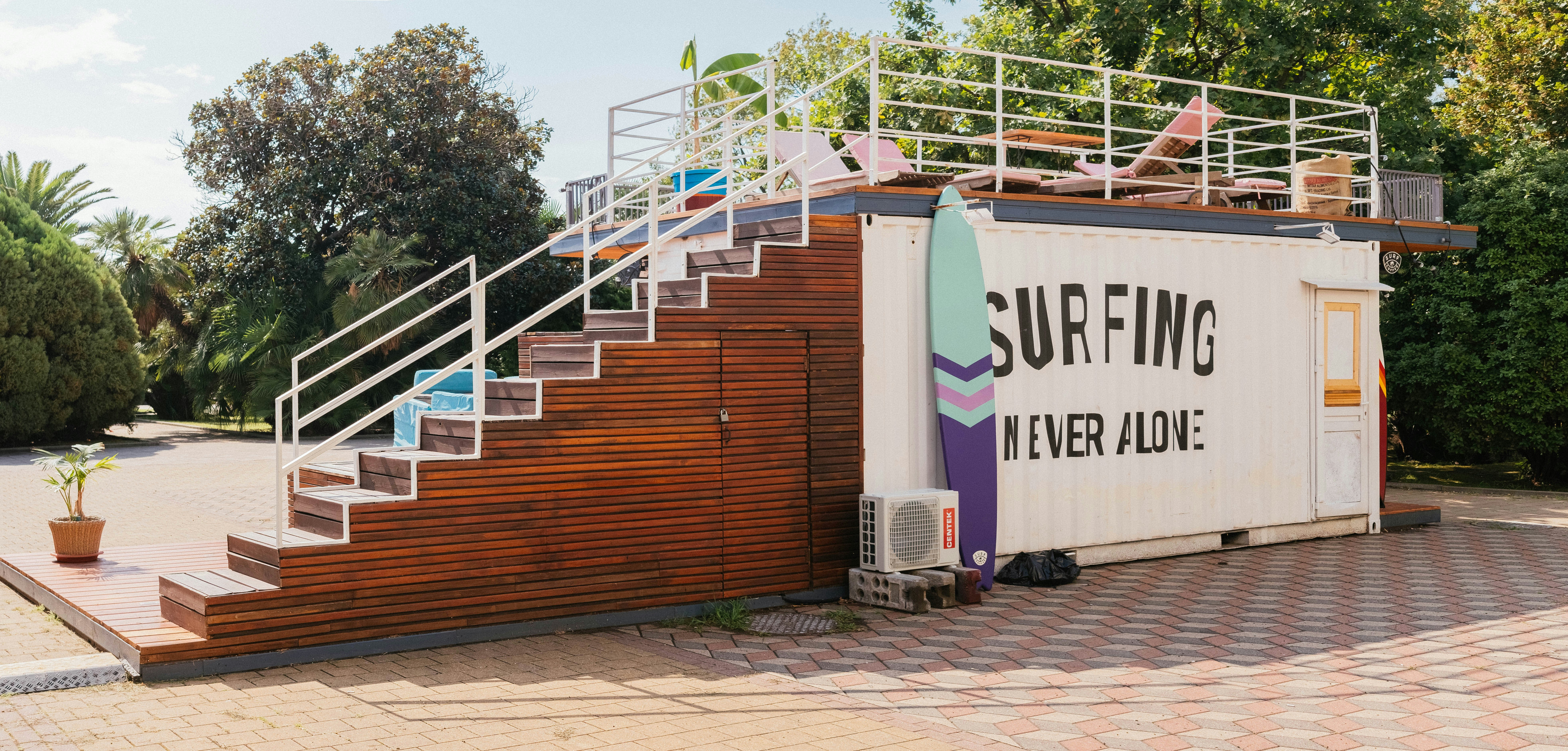 A container building is transformed into a surf-themed recreational area. It features a staircase made of wood leading up to a rooftop deck with a few chairs and surfboards as decoration. The side of the container has a large sign that reads 'Surfing Never Alone.' The area is surrounded by greenery, including trees and bushes, and the ground is paved with stone tiles.