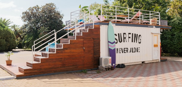 A container building is transformed into a surf-themed recreational area. It features a staircase made of wood leading up to a rooftop deck with a few chairs and surfboards as decoration. The side of the container has a large sign that reads 'Surfing Never Alone.' The area is surrounded by greenery, including trees and bushes, and the ground is paved with stone tiles.