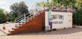 A container building is transformed into a surf-themed recreational area. It features a staircase made of wood leading up to a rooftop deck with a few chairs and surfboards as decoration. The side of the container has a large sign that reads 'Surfing Never Alone.' The area is surrounded by greenery, including trees and bushes, and the ground is paved with stone tiles.