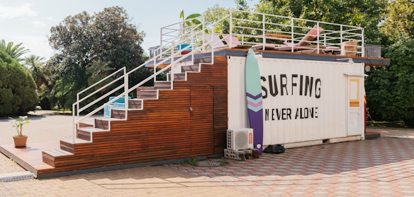 A container building is transformed into a surf-themed recreational area. It features a staircase made of wood leading up to a rooftop deck with a few chairs and surfboards as decoration. The side of the container has a large sign that reads 'Surfing Never Alone.' The area is surrounded by greenery, including trees and bushes, and the ground is paved with stone tiles.