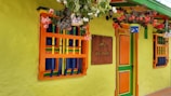 A brightly colored building facade featuring vivid green walls with orange and blue accents around the windows and doors. Potted plants with lush green leaves and red and white flowers hang above, adding to the vibrant and cheerful atmosphere. A sign on the wall reads 'Casa Hotel las Palmeras.'