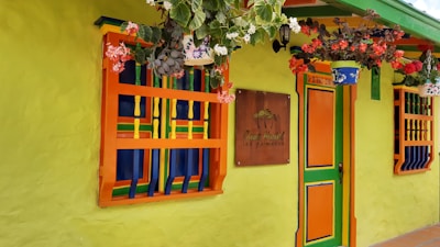 A brightly colored building facade featuring vivid green walls with orange and blue accents around the windows and doors. Potted plants with lush green leaves and red and white flowers hang above, adding to the vibrant and cheerful atmosphere. A sign on the wall reads 'Casa Hotel las Palmeras.'