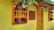 A brightly colored building facade featuring vivid green walls with orange and blue accents around the windows and doors. Potted plants with lush green leaves and red and white flowers hang above, adding to the vibrant and cheerful atmosphere. A sign on the wall reads 'Casa Hotel las Palmeras.'